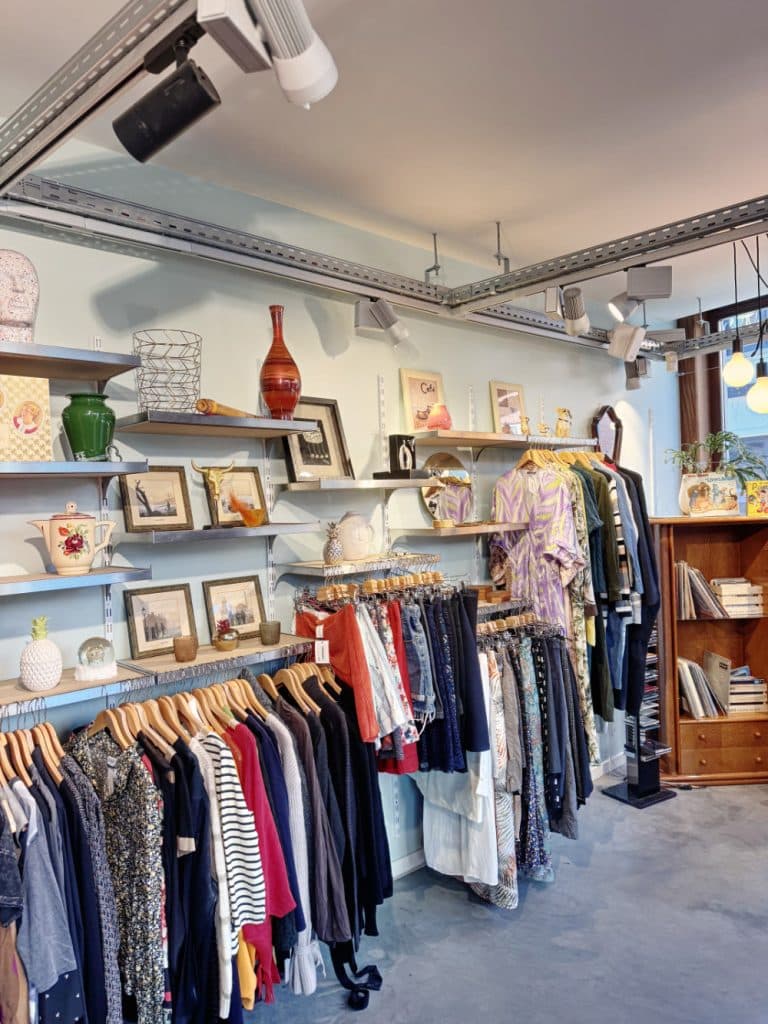 Kledingrekken met verschillende kleurrijke kledingstukken hangen onder planken met ingelijste foto's, kunst, vazen en decoratieve voorwerpen in een gezellige vintage winkelruimte in Hasselt, alles badend in zachte verlichting.