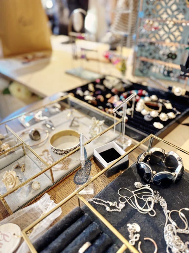 Een uitstalling van verschillende sieraden, zoals kettingen, ringen, armbanden, oorbellen en broches, in glazen bakjes en doosjes op een tafel in een helder verlichte vintage winkel in Hasselt.