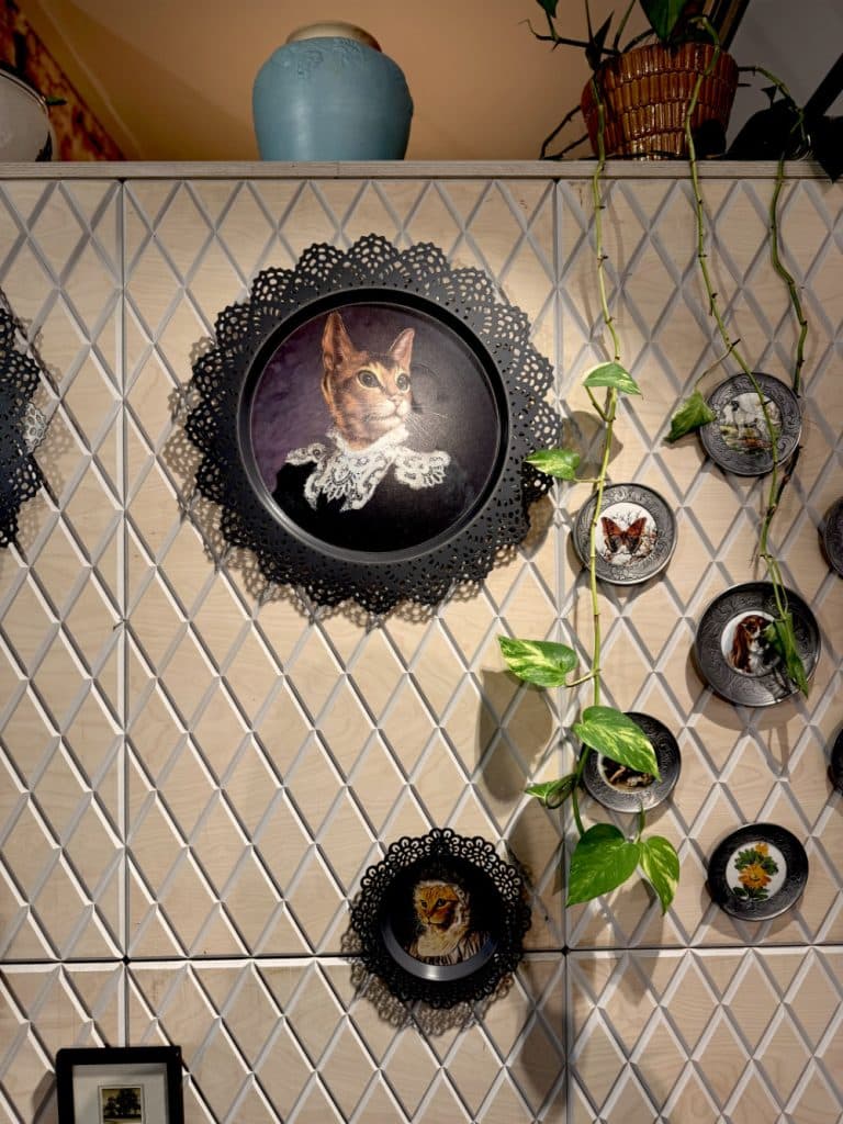 Een muur versierd met sierlijke ronde borden uit vintage shoppen in Hasselt heeft geschilderde portretten van katten en vlinders, een centraal bord met een kat in koninklijke kledij en een groene wingerd tussen de borden. Bovenop de muur staat een blauwe vaas.