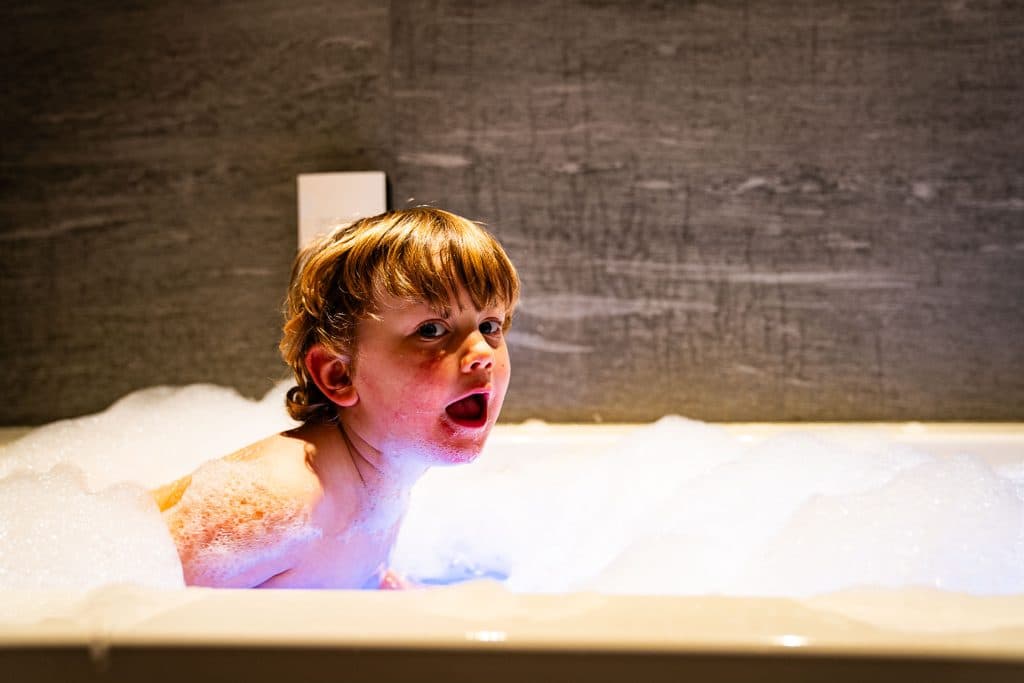 Een jong kind met nat haar en rode wangen zit in een bad vol bubbels en kijkt met open mond naar de camera. De achtergrond is een grijze betegelde muur, waardoor een gezellig tafereel ontstaat dat doet denken aan thuiskomen na een Pairi Daiza-bezoek.