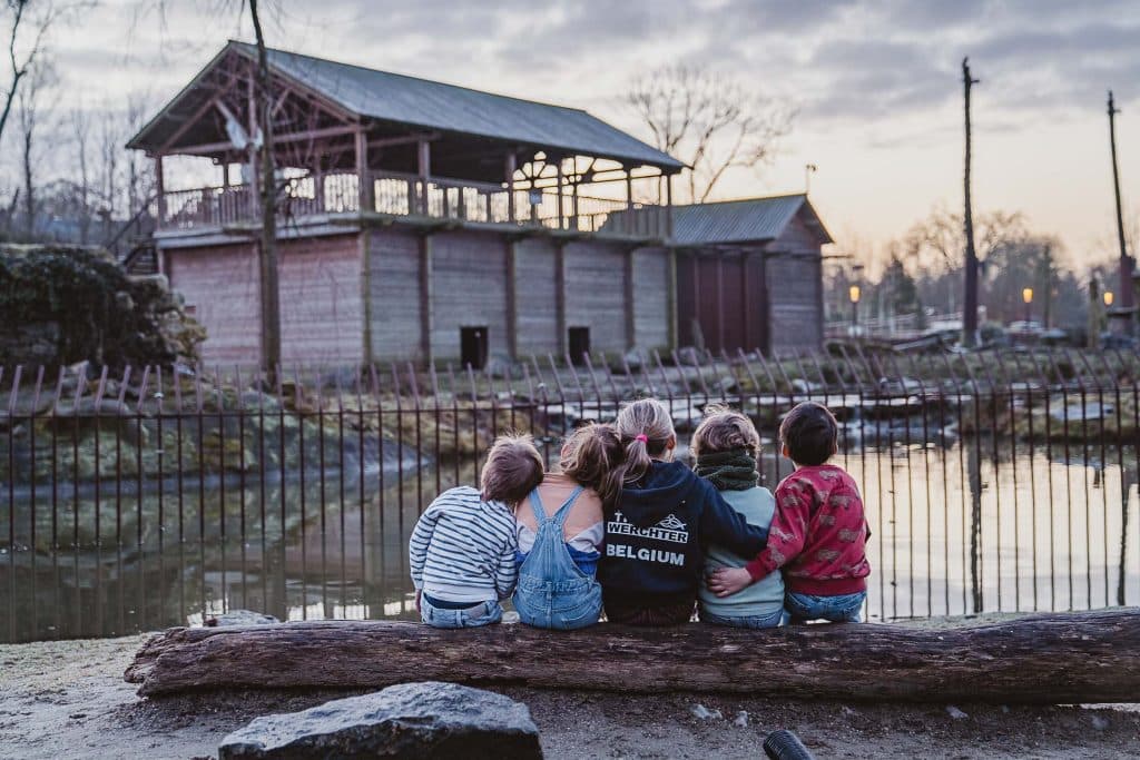 Vijf kinderen zitten op een boomstam bij een vijver in Pairi Daiza, armen om elkaar heen, uitkijkend op rustieke gebouwen onder een bewolkte avondhemel. De sfeer is vredig en reflectief - perfect voor wie houdt van een dierentuinbezoek met vrienden.