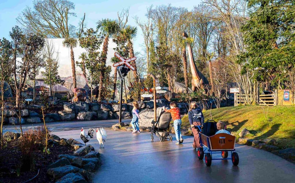 Kinderen en volwassenen lopen over een dierentuinpad met kinderwagens en een bolderkar, langs een dinosaurustentoonstelling met levensgrote modellen en een spoorwegovergang, omringd door bomen en rotsen tijdens hun Pairi Daiza bezoeken.