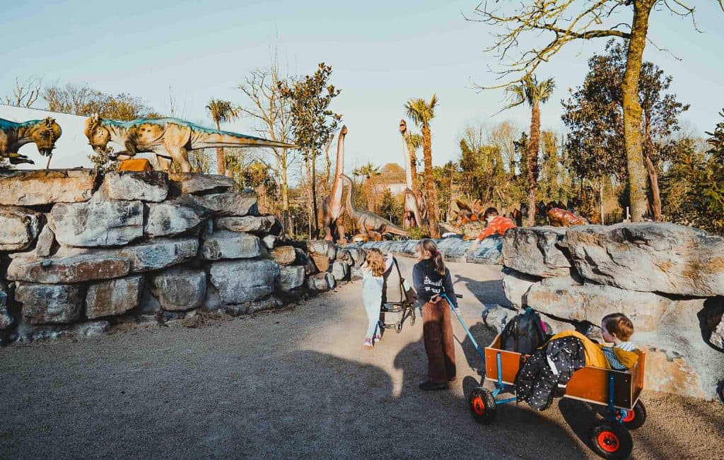 Twee kinderen trekken een kar met een jong kind erin over een pad omringd door grote dinosaurusbeelden en rotsen in Pairi Daiza dierentuin op een zonnige dag.
