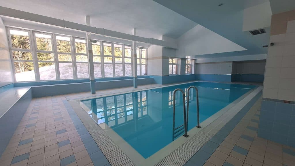 Een helder binnenzwembad met blauw betegelde randen, een metalen zwembadtrap en grote ramen die natuurlijk licht binnenlaten en een besneeuwd landschap buiten tonen - perfect na een dag skiën in Tsjechie. Het zwembad is schoon en ruim.