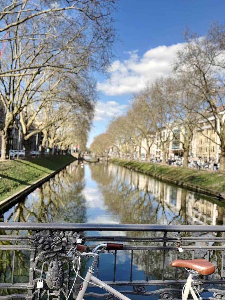 Een witte fiets staat geparkeerd op een brug in Düsseldorf, met uitzicht op een met bomen omzoomd kanaal met weerspiegelingen van bomen en gebouwen in het kalme water onder een blauwe lucht met verspreide wolken - perfect voor fietsen door Düsseldorf.