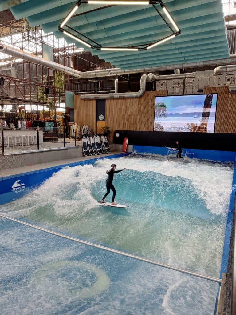 Een persoon surft op een kunstmatige golf in een indoor surfcentrum in Düsseldorf, met een andere persoon vlakbij. De locatie heeft een industrieel decor, zitplaatsen en een groot scherm - perfect om te ontspannen na het fietsen door Düsseldorf.