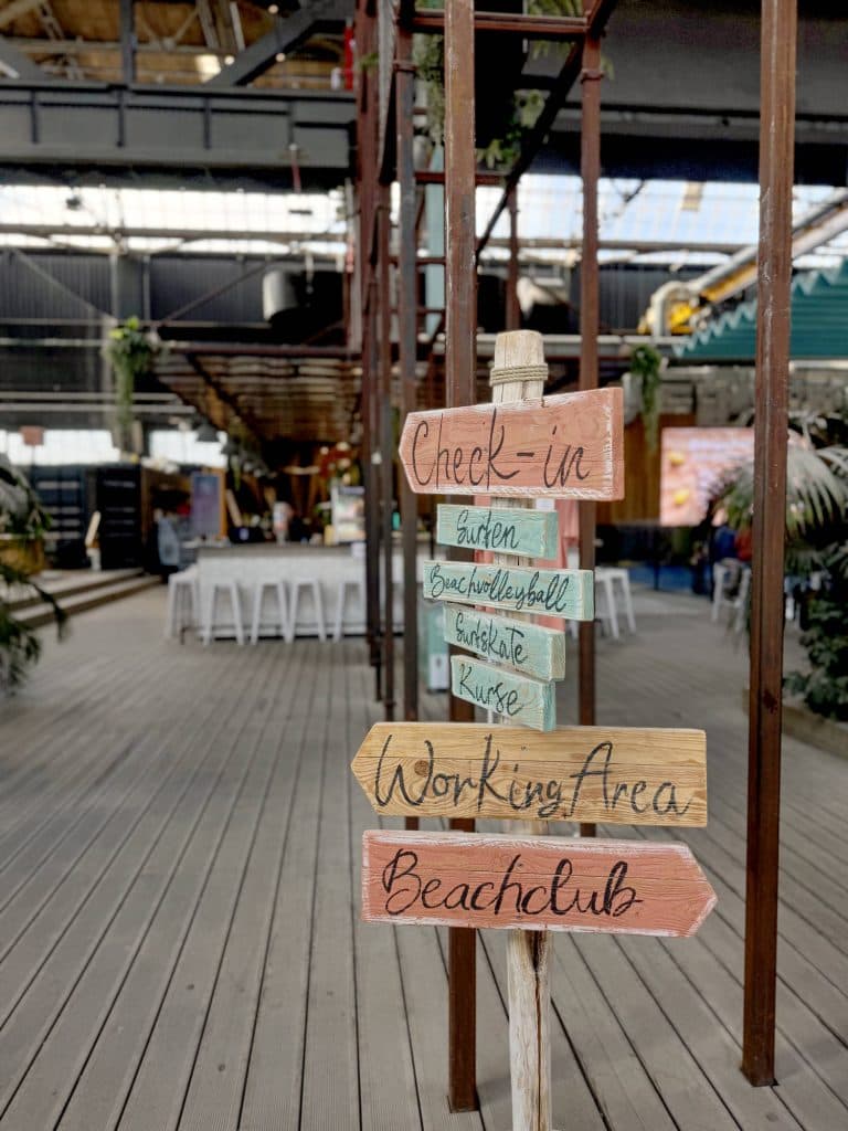 Kleurrijke houten wegwijzers binnen wijzen naar "Inchecken", "Surfen" en meer, met tafels en planten op de achtergrond die gasten uitnodigen om op ontdekkingstocht te gaan zoals fietsen door Düsseldorf, Duitsland.