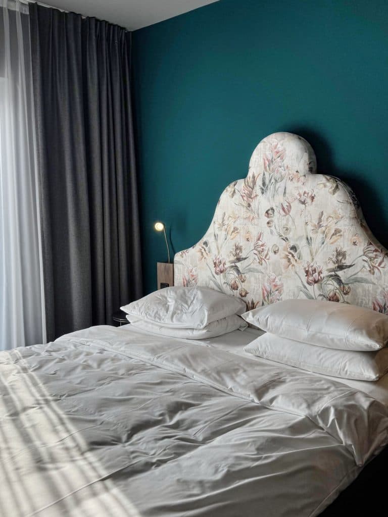 Een keurig opgemaakt bed met wit linnengoed en vier kussens staat tegen een groenblauwe muur, die de charme van Düsseldorf oproept. Het bed heeft een gestoffeerd hoofdeinde met bloemmotief, met daglicht dat door donkere gordijnen naar binnen valt en een klein bedlampje dat brandt.