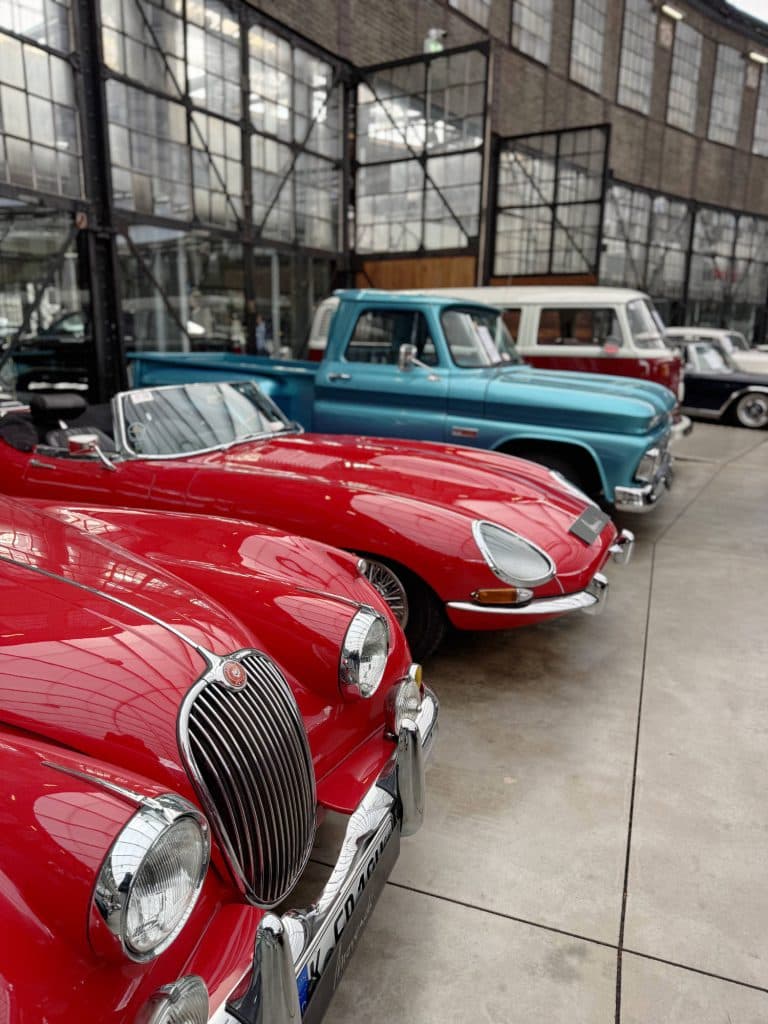 Een rij klassieke auto's in een garage in Düsseldorf, waaronder een glimmende rode cabriolet op de voorgrond en een vintage blauwe pick-up truck verder naar achteren geparkeerd bij grote industriële ramen. Perfect om te spotten tijdens het fietsen door Düsseldorf.