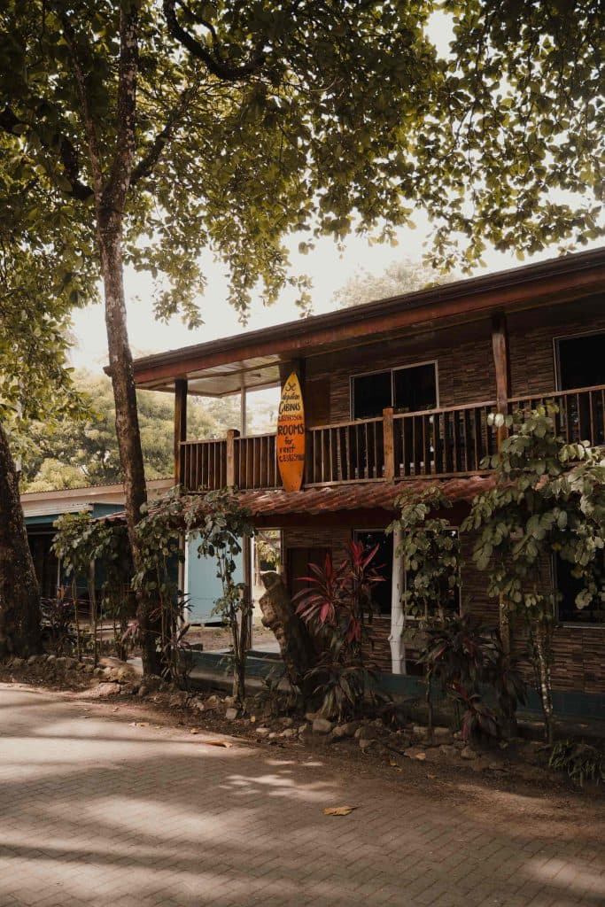 Een rustiek houten gebouw met een balkon wordt omringd door weelderige groene bomen en planten - perfect voor een vakantie in Costa Rica. Een gele surfplank met tekst leunt tegen de reling terwijl het zonlicht door de bladeren filtert en schaduwen werpt op het tafereel.