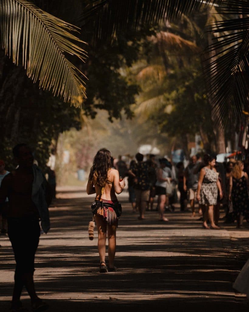 Een vrouw in een kleurrijke rok loopt door een zonovergoten, met palmbomen omzoomde straat vol mensen - een levendige, tropische scène die de levendige geest van vakantie in Costa Rica weergeeft, waar het gedempte zonlicht door de bomen schijnt.