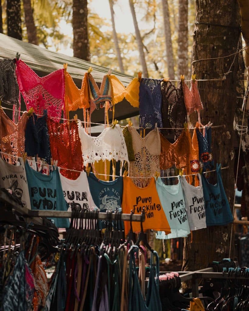 Kleurrijke gehaakte topjes en grafische draagtassen met zinnen als "Pura Vida" en "Pura Fucking Vida" hangen uitgestald op een openluchtmarkt in Costa Rica, met palmbomen en zonlicht die de geest van wat doen in Costa Rica vastleggen.