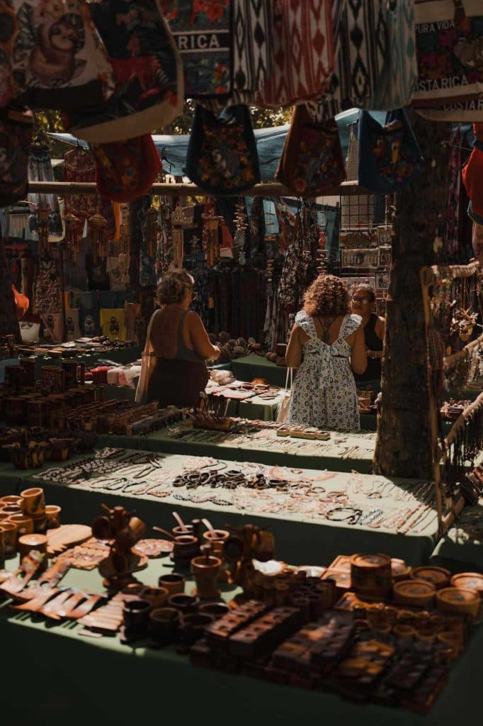 Een levendige markt in de open lucht met tafels met kleurrijke ambachten, sieraden en houten voorwerpen - net als op de vakantie in Costa Rica - waar drie mensen snuffelen onder hangend textiel en souvenirs in warm zonlicht en diepe schaduwen.