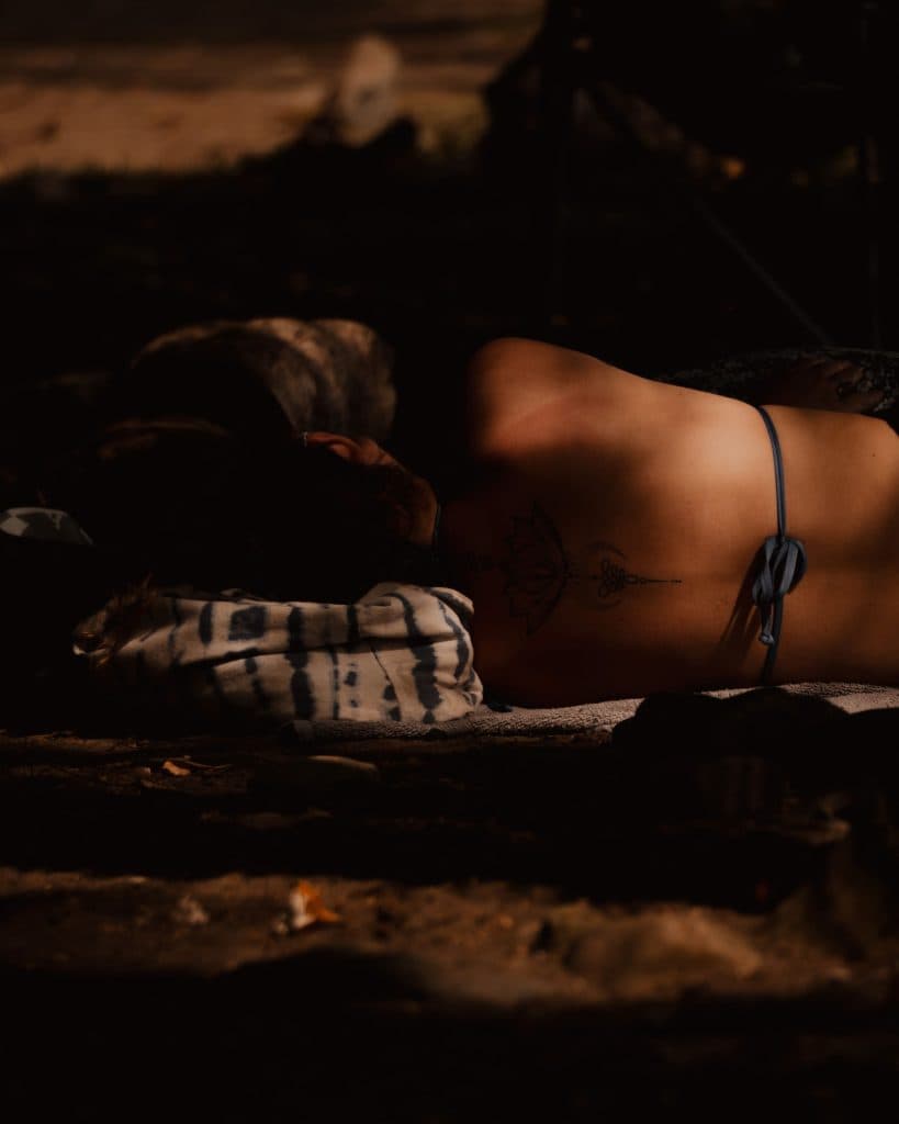 Een persoon ligt buiten op zijn buik in Costa Rica, rustend op een kussen met een patroon. Hun rug is gedeeltelijk ontbloot en onthult een tatoeage en een blauw bikinibandje, met schemerig licht en zacht zonlicht dat door de bomen van het nationale park schijnt.