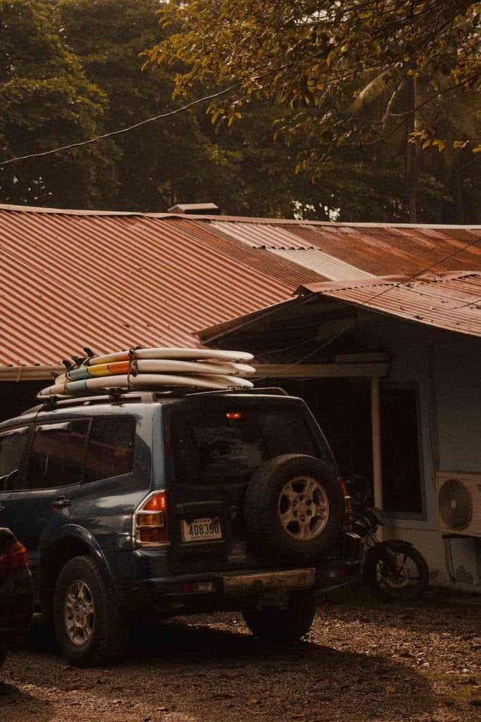 Een blauwe SUV met een reservewiel en verschillende surfplanken op het dak staat geparkeerd naast een gebouw met een verroest metalen dak, omringd door bomen en groen - perfect voor een avontuurlijke vakantie in Costa Rica.