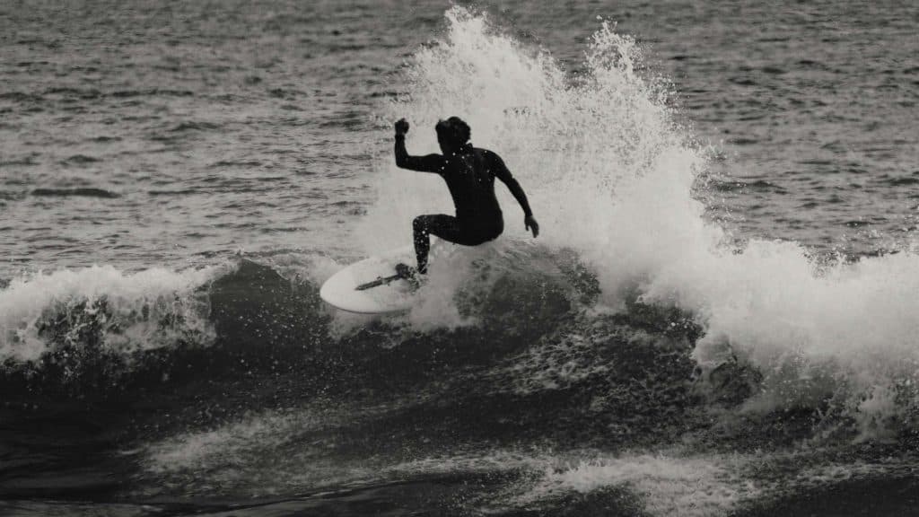 Een surfer in een wetsuit berijdt een brekende golf, midden in actie vastgelegd met dramatisch opspattend water achter hem. Deze zwart-wit scène toont de sensatie van surfen - een van de leukste dingen om te doen tijdens een vakantie in Costa Rica.