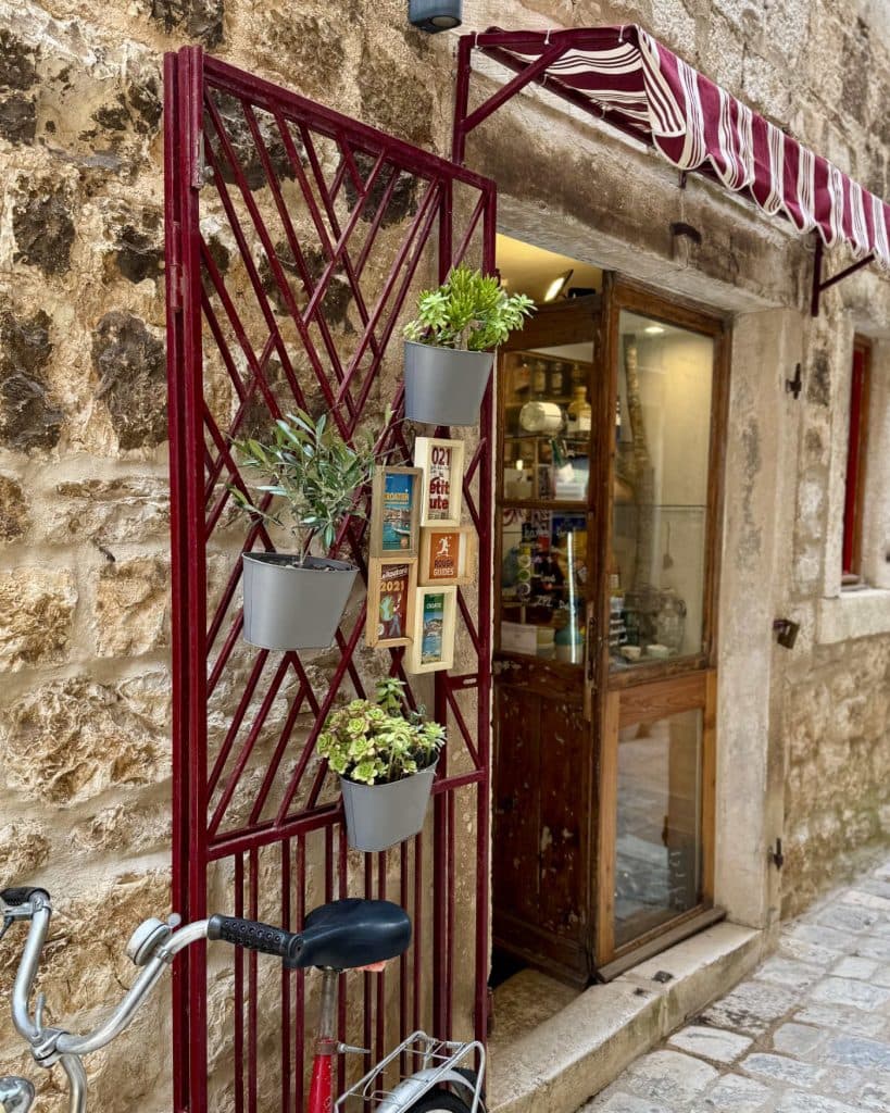 Een rood metalen hek met potplanten hangt naast de ingang van een stenen gebouw in Hvar, Kroatie. Boven de houten deur hangt een rood-wit gestreepte luifel en binnen zijn planken met keramiek te zien. Vlakbij staat een fiets geparkeerd.