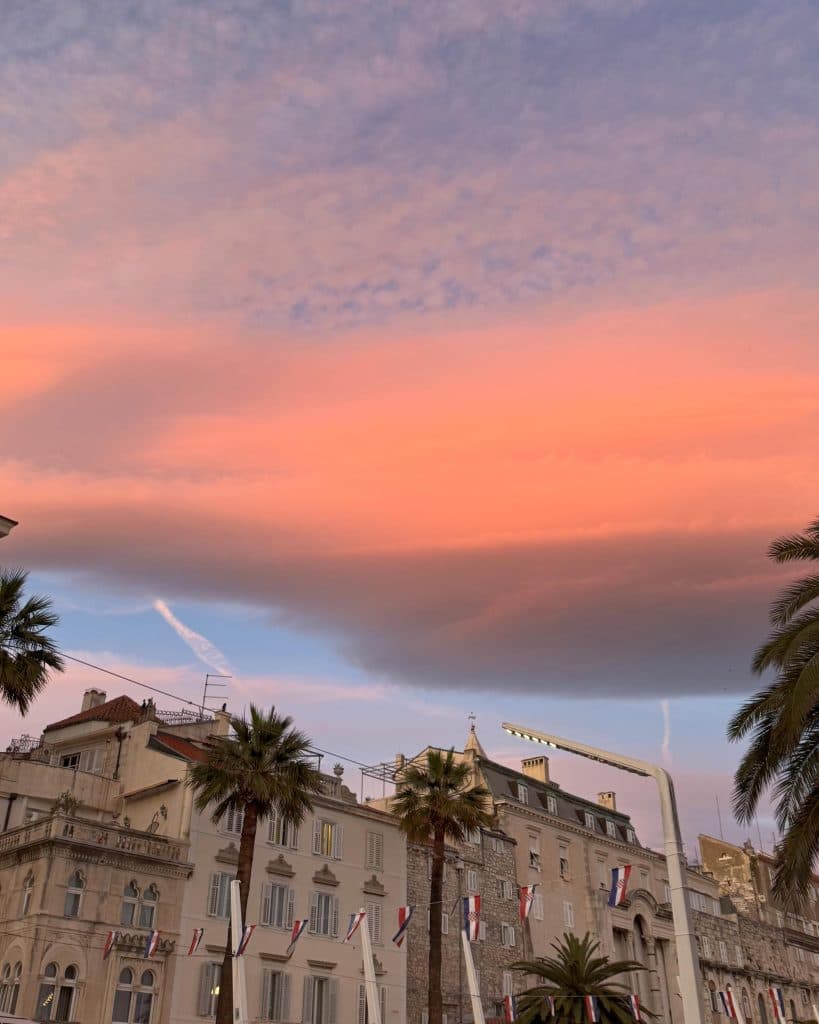 Een schilderachtig uitzicht op pastelkleurige gebouwen en palmbomen onder een levendige hemel met roze en oranje wolken bij zonsondergang. Split in Kroatie
