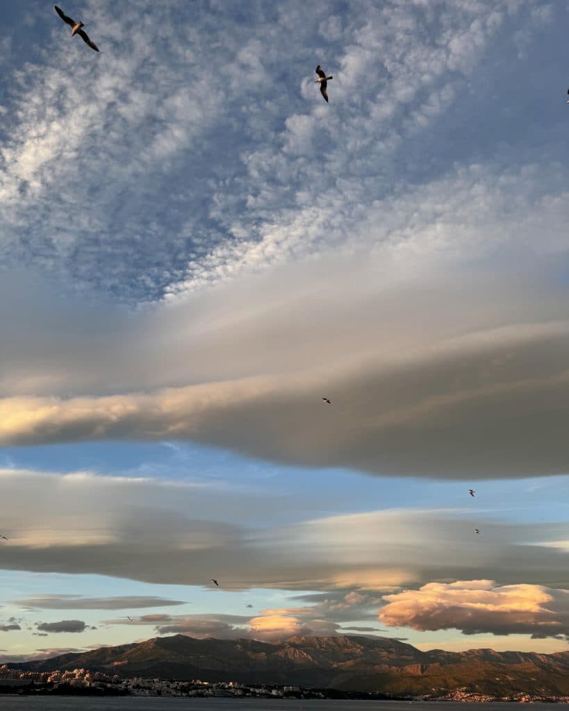 Gouden wolken strekken zich uit over een blauwe hemel bij zonsondergang boven Loonseche Land, waar vogels over verre bergen zweven en het betoverende stadje de Efteling aan de horizon glinstert - een perfect tafereel voor een vakantie met kinderen.