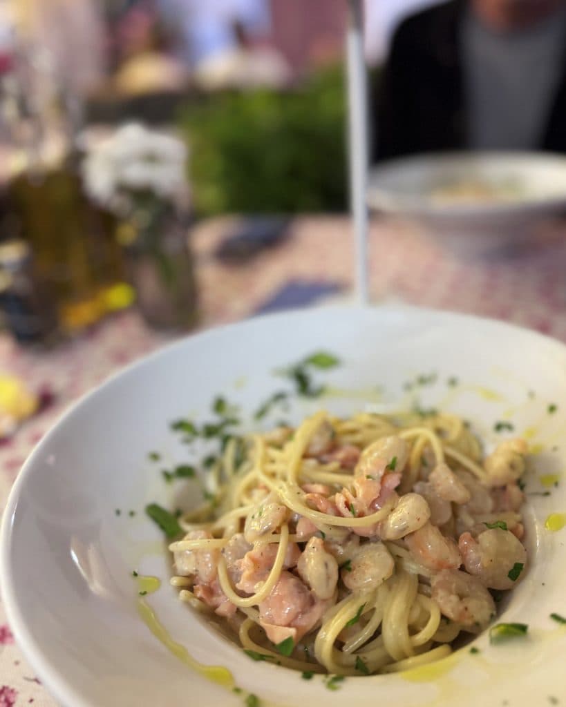 Een close-up van een bord spaghetti met garnalen en kruiden, besprenkeld met olijfolie. De zacht vervaagde achtergrond verwijst naar eilandhoppen in Kroatië, met flessen en bloemen die een ontspannen mediterrane sfeer oproepen.