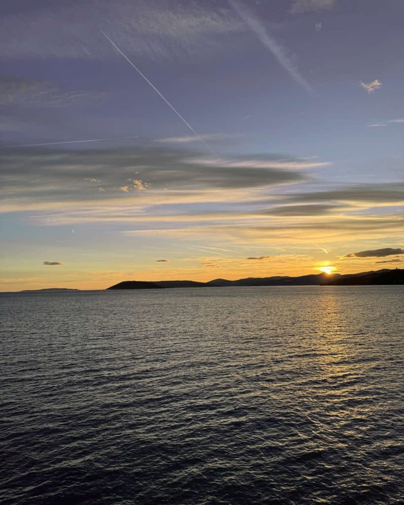 Zonsondergang over een kalme zee met zachte rimpelingen, heuvels in silhouetten aan de horizon en wolkenstrepen in een kleurrijke lucht die overgaat van blauw naar oranje - perfect voor eilandhoppen of waterval spotten in Kroatië.