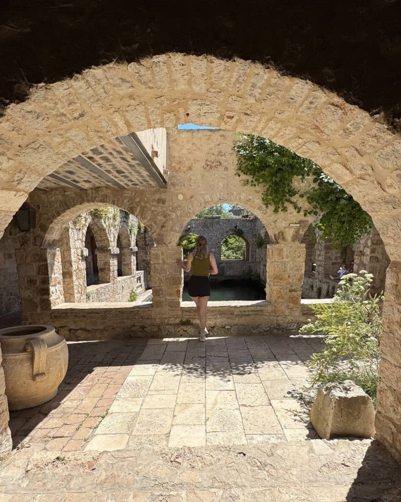 Een persoon met een rugzak loopt door een zonovergoten stenen boog in een oude binnenplaats in Kroatië, omringd door bogen, planten en een grote kleipot aan de linkerkant. Helder licht benadrukt de historische architectuur - perfect voor eilandhoppen of waterval spotten in de buurt.