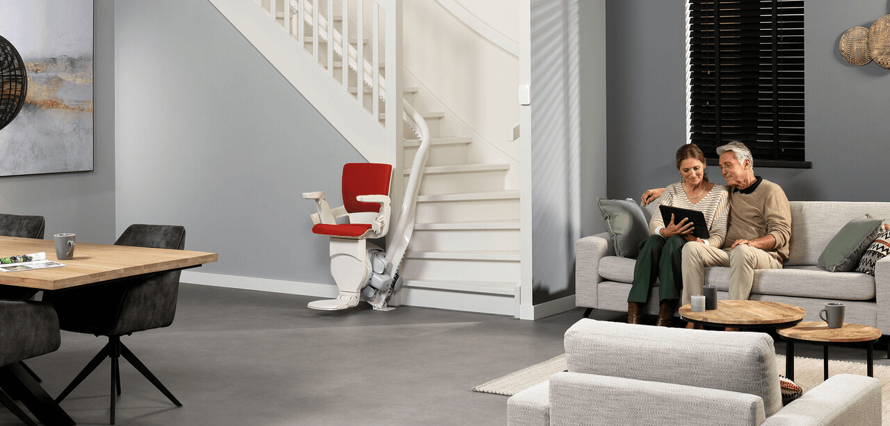 Een stel zit op een bank en bekijkt een fotoalbum in hun moderne vakantiehuis. Op de achtergrond maakt een rood-witte traplift aan de trap de lichte, neutraal getinte ruimte klaar voor de toekomst.