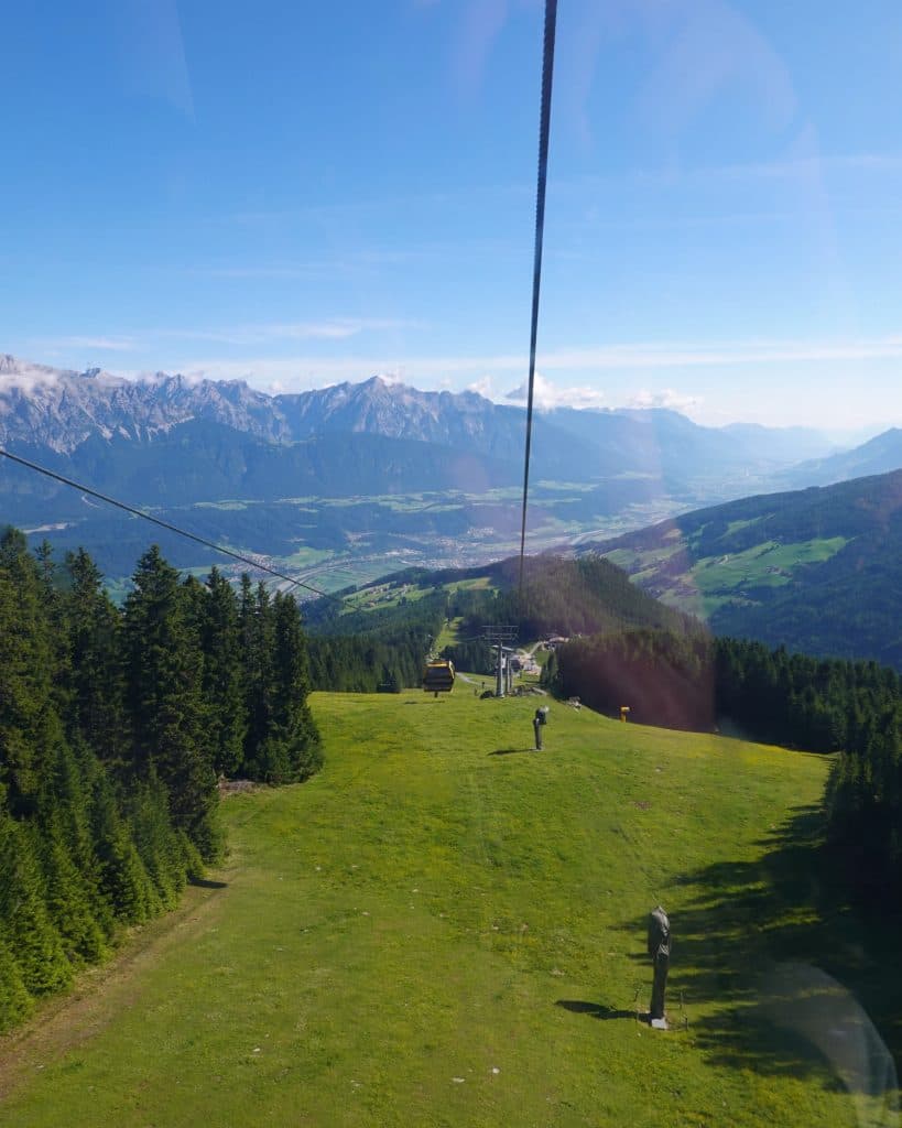 Een schilderachtig uitzicht vanuit een kabelbaan omhoog over een groene berghelling met dennenbomen, uitkijkend over een vallei met bergen in de verte onder een strakblauwe hemel.