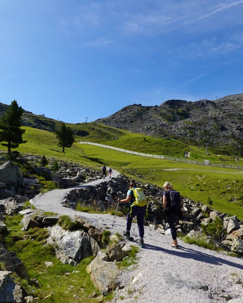 Wandelaars lopen op een zonnige dag over een rotsachtig pad, omgeven door groene heuvels en verspreide bomen, met een blauwe lucht boven hun hoofd en bergen op de achtergrond.