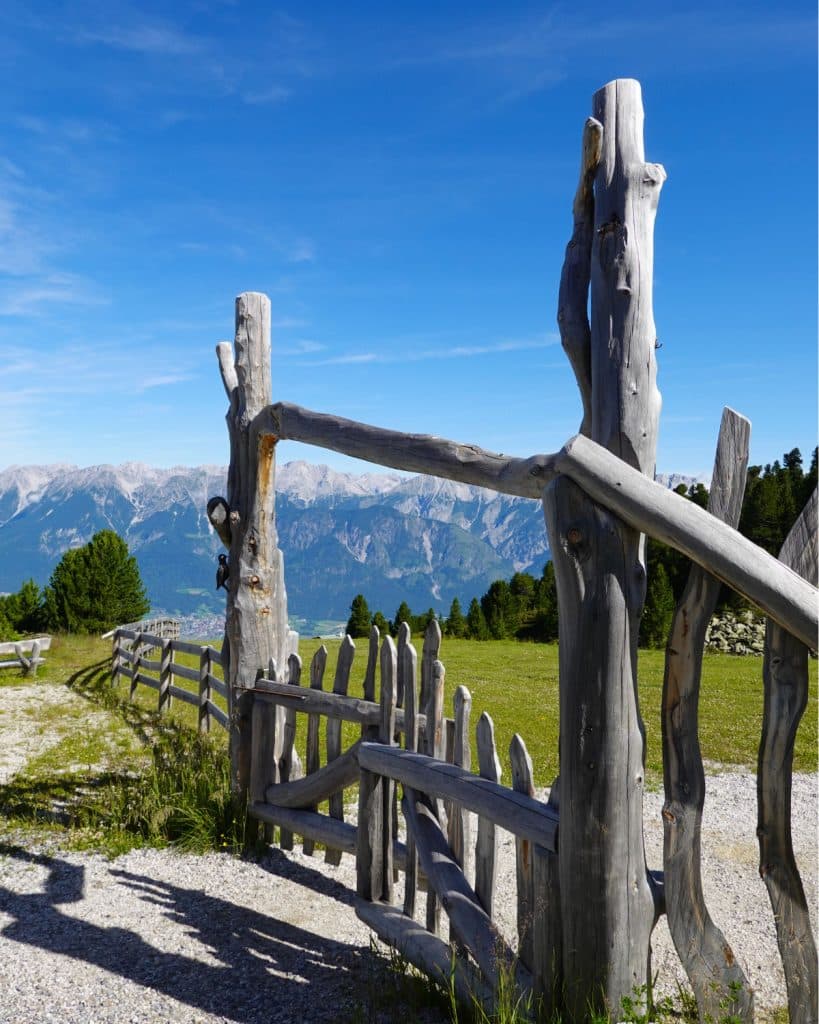Een rustiek houten hek en poort staan open op een grindpad, met groene velden, bomen en bergen in de verte onder een helderblauwe hemel op de achtergrond.