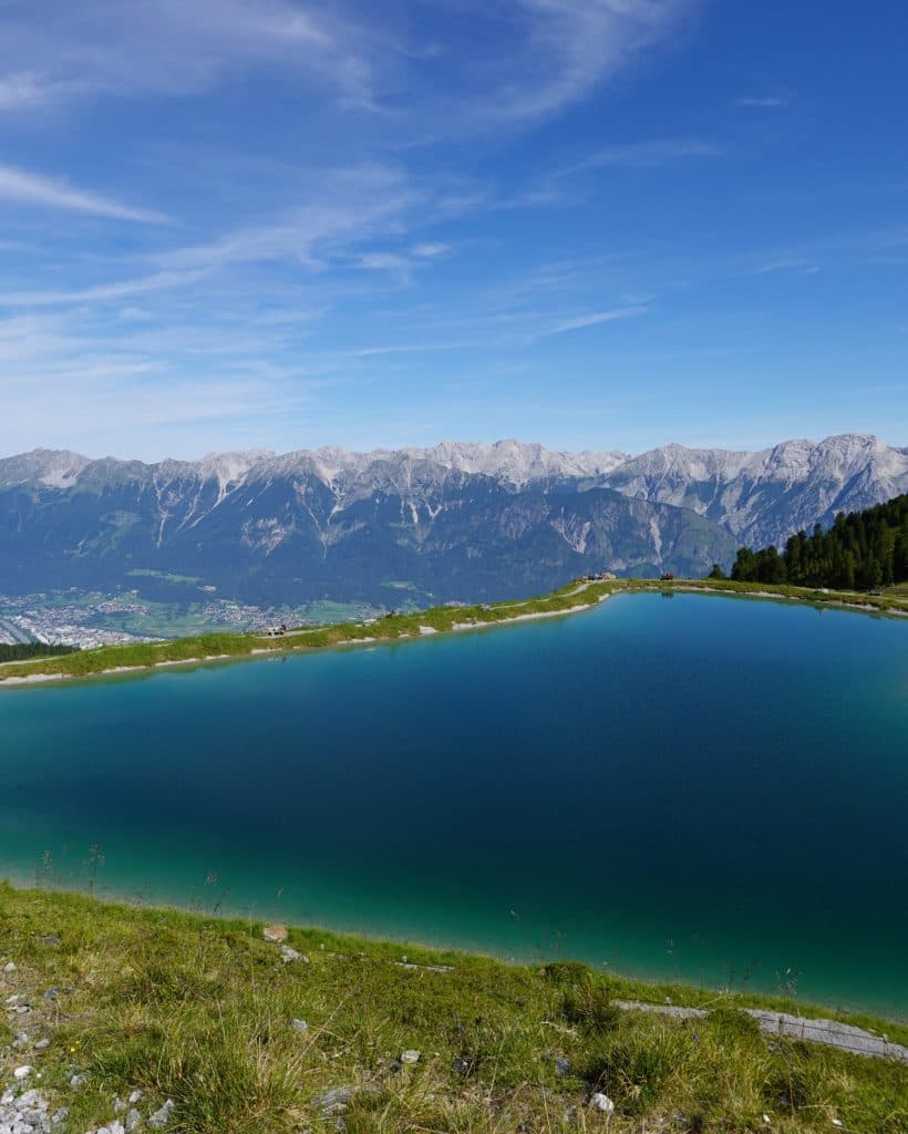 Een helderblauw bergmeer omzoomd door groen gras, met in de verte besneeuwde bergen en een blauwe lucht met pluizige wolken op de achtergrond.