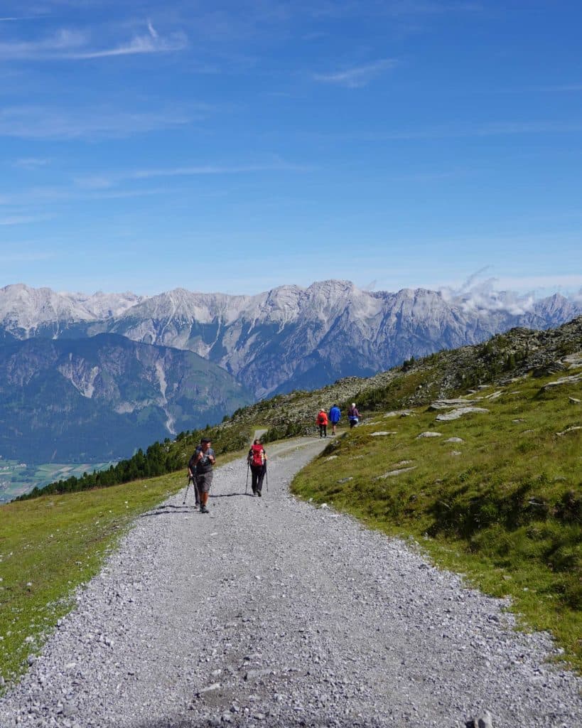Een groep wandelaars loopt over een grindpad op een met gras begroeide berghelling, met ruige bergtoppen en een helderblauwe lucht op de achtergrond.
