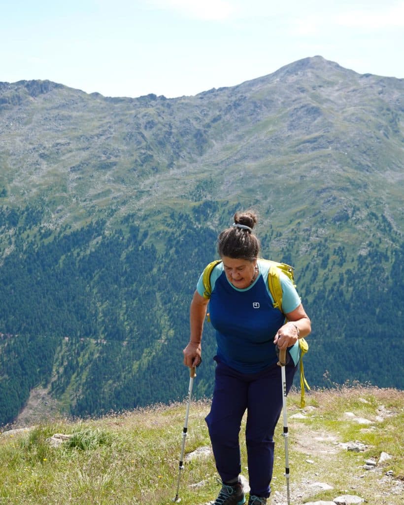 Een vrouw met wandelstokken loopt een bergpad op, draagt een blauw shirt, een donkere broek en een gele rugzak, met groene heuvels en bergtoppen op de achtergrond onder een heldere hemel.