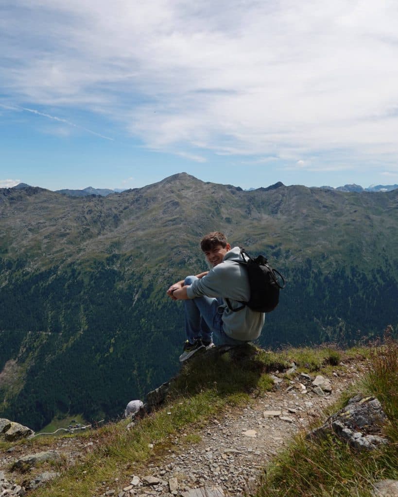 Een persoon met een rugzak zit aan de rand van een bergpad, glimlacht naar de camera, met een schilderachtig uitzicht op groene valleien en ruige bergtoppen onder een gedeeltelijk bewolkte hemel op de achtergrond.