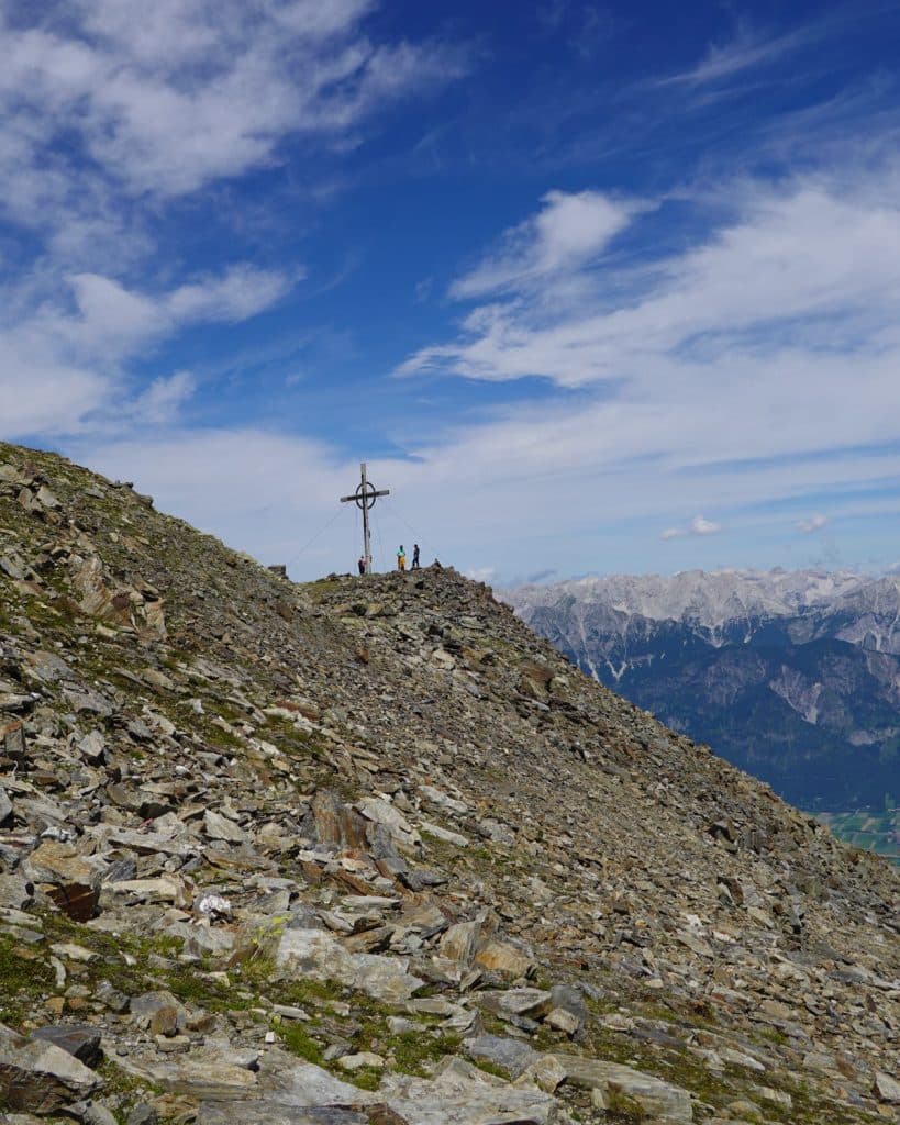 Een rotsachtige berghelling met een groep mensen bij een hoog kruis op de top, onder een gedeeltelijk bewolkte blauwe lucht en met ruige bergen in de verte op de achtergrond.