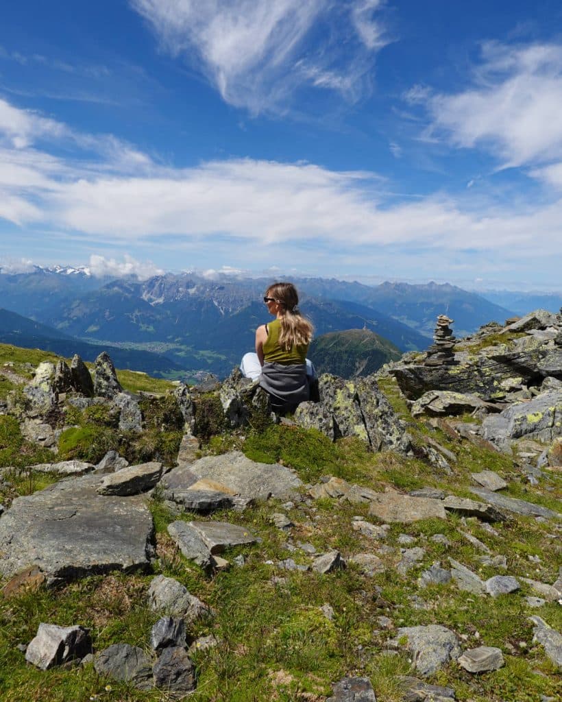 Een vrouw zit op een rotsachtig terrein en kijkt weg, met uitzicht op een schilderachtig berglandschap onder een blauwe hemel met pluizige wolken. Gestapelde stenen en stukjes groen gras omringen haar.
