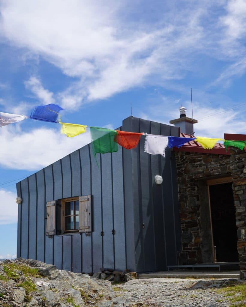 Een klein stenen en metalen gebouw met houten luiken staat onder een blauwe hemel. Boven de ingang hangen kleurrijke gebedsvlaggen die wapperen in de wind. Rotsachtige grond omringt het gebouwtje.