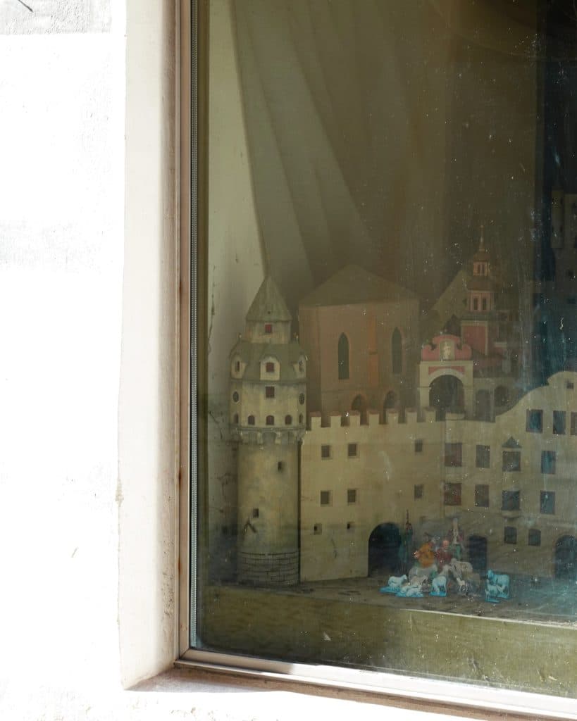 Een modelkasteel met torens en muren staat achter een glazen raam, dat het licht gedeeltelijk weerkaatst. De scène is geïnspireerd op Tirol en bevat miniatuurgebouwen en kleine figuurtjes op de grond - perfect voor je zomerchecklist van dingen die je moet doen.