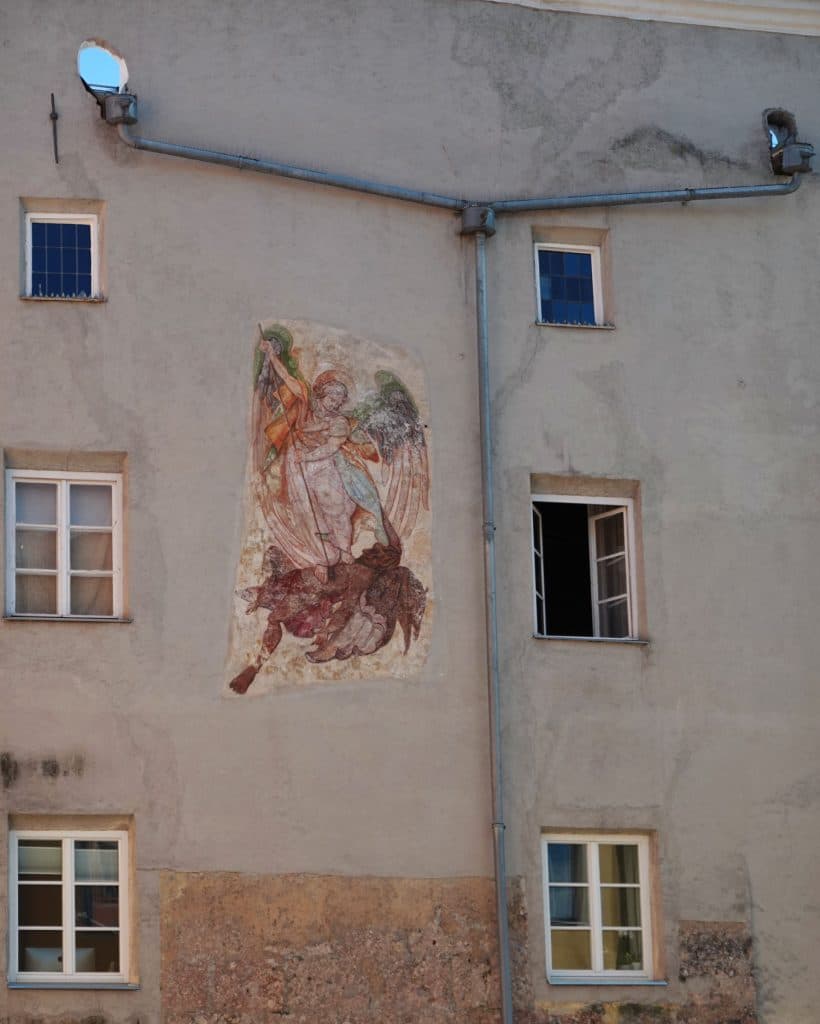 Een vervaagde muurschildering van een engel die met een draak vecht siert de beige gevel van een oud gebouw in Tirol, tussen drie gesloten ramen en één open raam. Grijze afvoerbuizen lopen langs de muur - een unieke plek om toe te voegen aan je Zomerchecklist van dingen om te doen in Tirol.