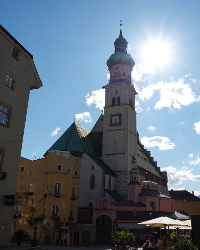 Een hoge kerk met een klokkentoren staat tegen een helderblauwe lucht in Tirol, de zon schijnt erboven. Op de voorgrond zie je verschillende kleurrijke gebouwen en zitjes met mensen die van hun tijd genieten - een perfecte plek op je Zomerchecklist.