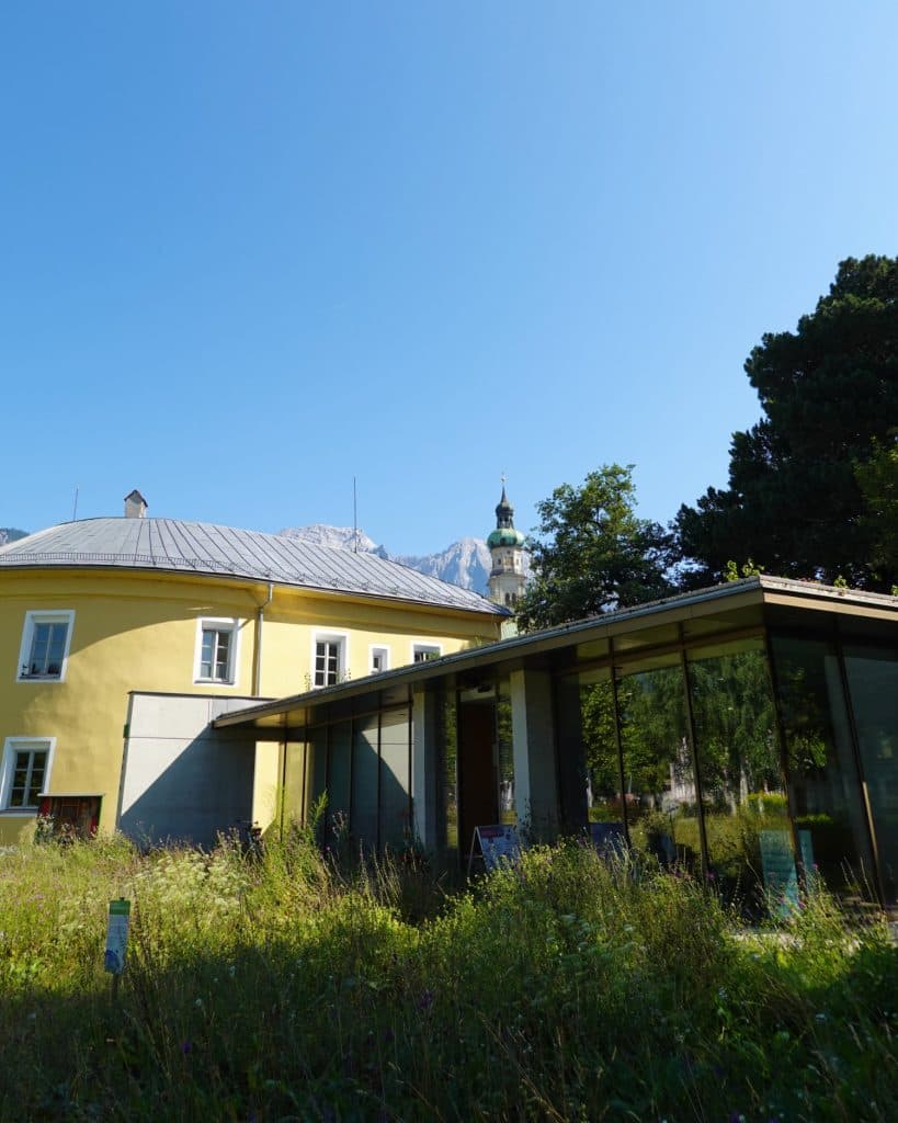 Een geel gebouw met een grijs dak staat naast een modern glazen bouwwerk in Tirol, omringd door hoog gras en bomen onder een strakblauwe hemel, met een kerktoren en bergen zichtbaar op de achtergrond.