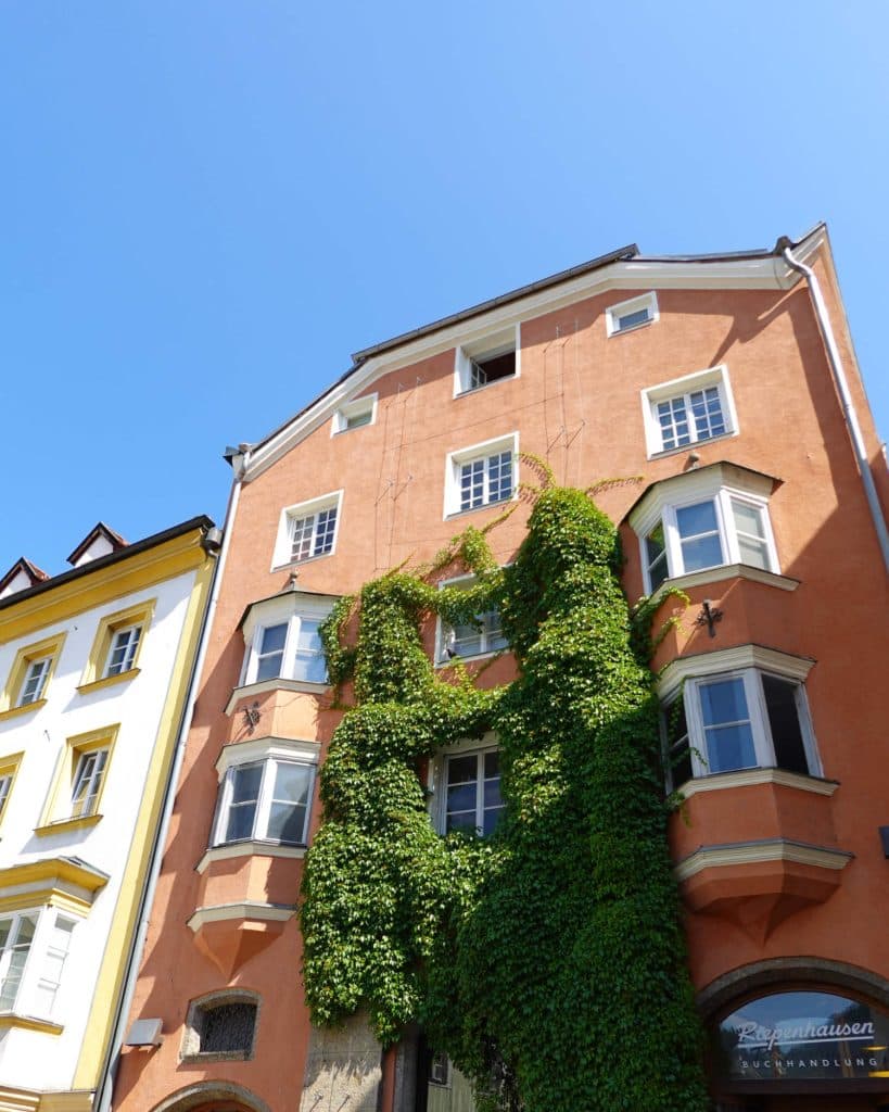 Een hoog, oranje gebouw met wit omrande ramen is bedekt met weelderig groene klimop. Het staat naast een geel gebouw onder een strakblauwe hemel - het perfecte decor voor je zomerchecklist in Tirol.