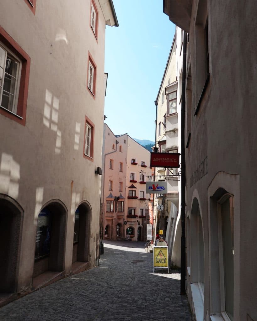 Een smalle straat met kinderkopjes, omzoomd door hoge, oude gebouwen met gewelfde deuropeningen en roodomrande ramen, die leidt naar een zonnig plein - perfect voor je Tirol zomerchecklist wandeling tussen pastelkleurige gebouwen in de verte.