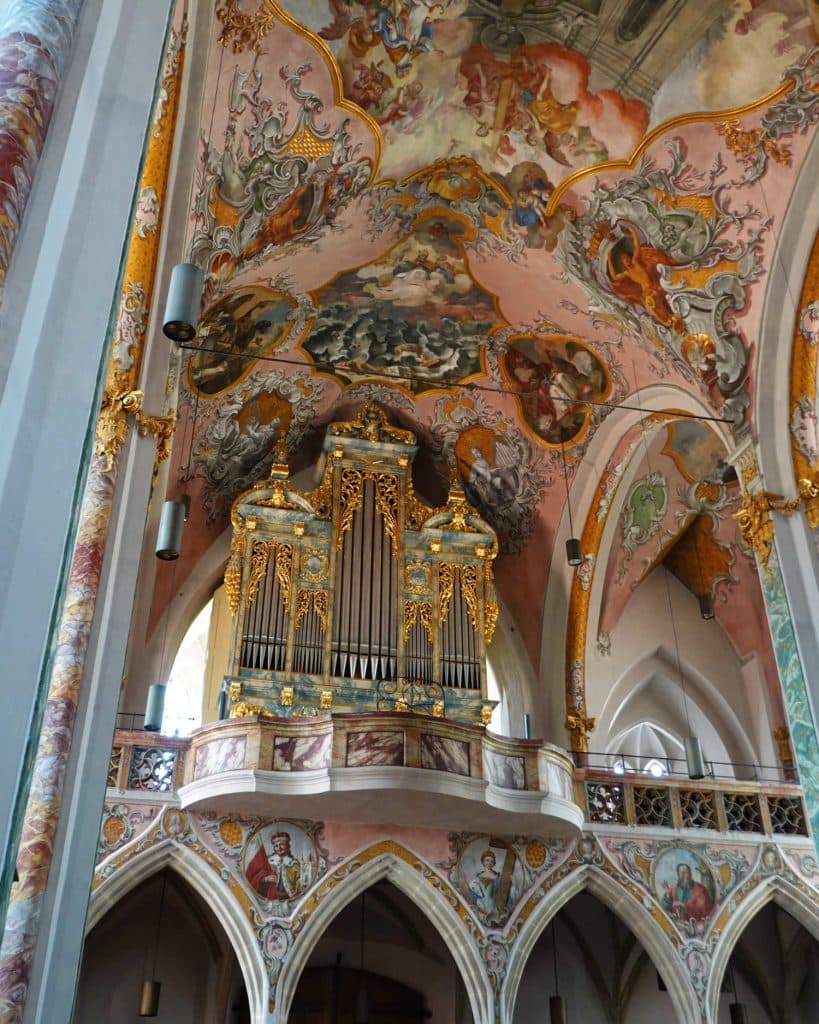 Een rijkelijk versierd kerkinterieur in Tirol heeft een sierlijk pijporgel met gouden details, onder een gewelfd plafond versierd met kleurrijke, uitgebreide fresco's en ingewikkelde kunstwerken. Boogramen en zuilen omlijsten het levendige tafereel.