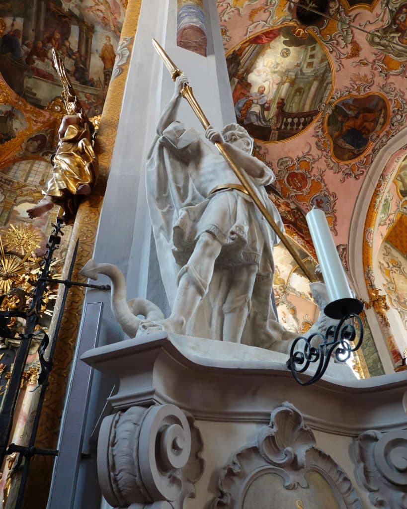 Een marmeren beeld van een man in harnas die een speer boven een slang houdt, waarschijnlijk Sint Joris voorstellend, staat in een sierlijke kerk in Tirol met fresco's op het plafond en decoratieve gouden accenten-een waar hoogtepunt voor elke Zomerchecklist.