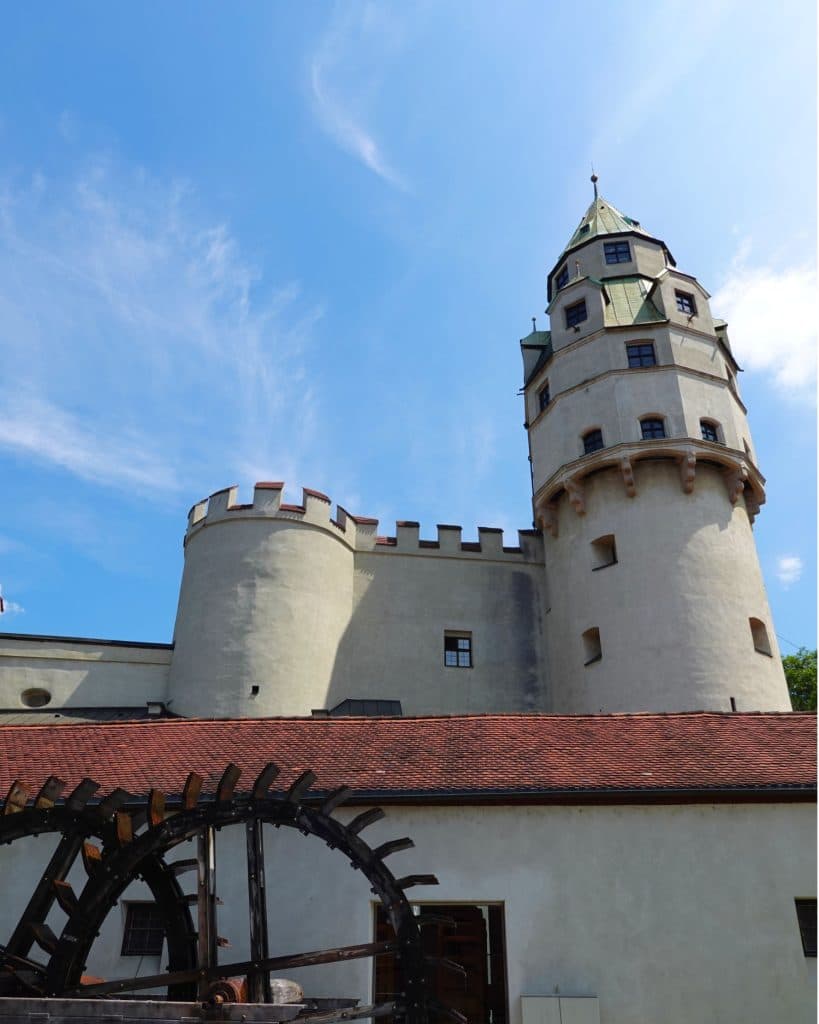 Een stenen kasteel met een ronde toren en kegelvormig dak rijst op achter een rood betegeld gebouw in Tirol. Een groot houten waterrad is zichtbaar op de voorgrond onder een helderblauwe hemel, perfect voor je Zomerchecklist.