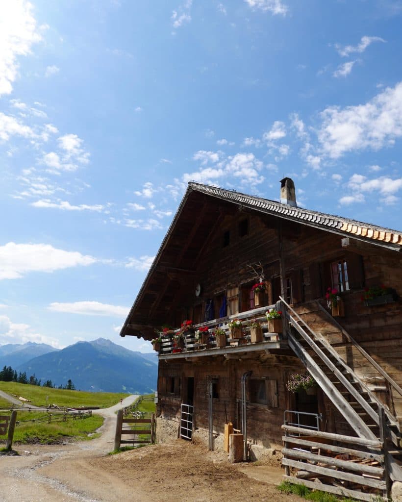 Een rustiek houten huis met bloembakken op een balkon staat naast een zandpad in Tirol, omringd door groene velden en bergen onder een blauwe hemel - de perfecte scène voor je Zomerchecklist-avontuur.