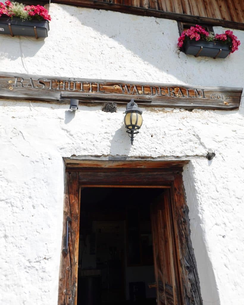 Houten ingang van een rustiek wit gebouw in Tirol met bloembakken en een lantaarn boven de deur. Op een houten bordje staat "Rasthütte Walderalm 1501". De open deur onthult een schemerig interieur - perfect voor je zomerchecklist van dingen die je moet doen.