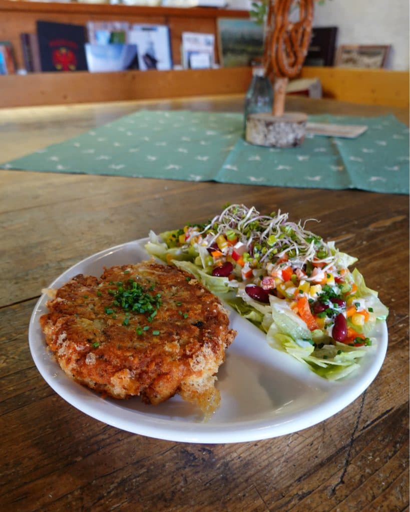 Een wit bord met een krokant gepaneerde patty gegarneerd met kruiden en een kleurrijke salade van sla, maïs, bonen, spruiten en groenten staat op een houten tafel - perfect voor je zomerchecklist van dingen die je moet doen in gezellig, rustiek Tirol.