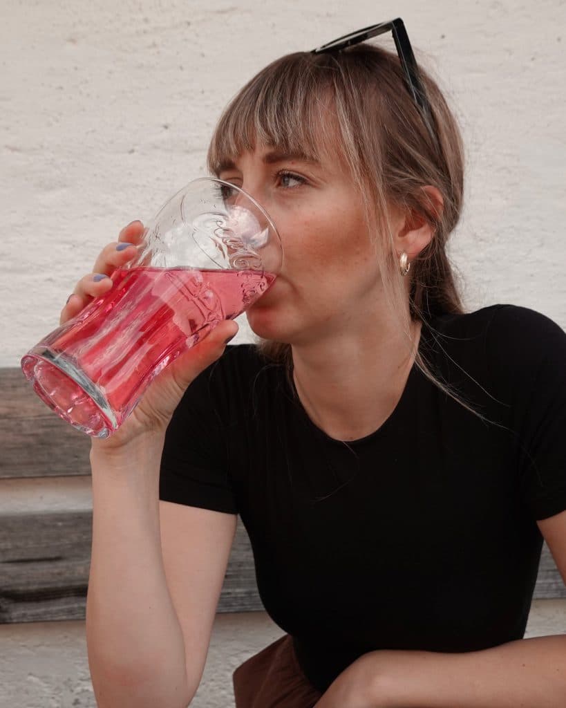 Een vrouw met lichtbruin haar en pony, een zwart shirt en een zonnebril op haar hoofd, zit op een bankje in Tirol en drinkt een roze drankje uit een Coca-Cola glas.